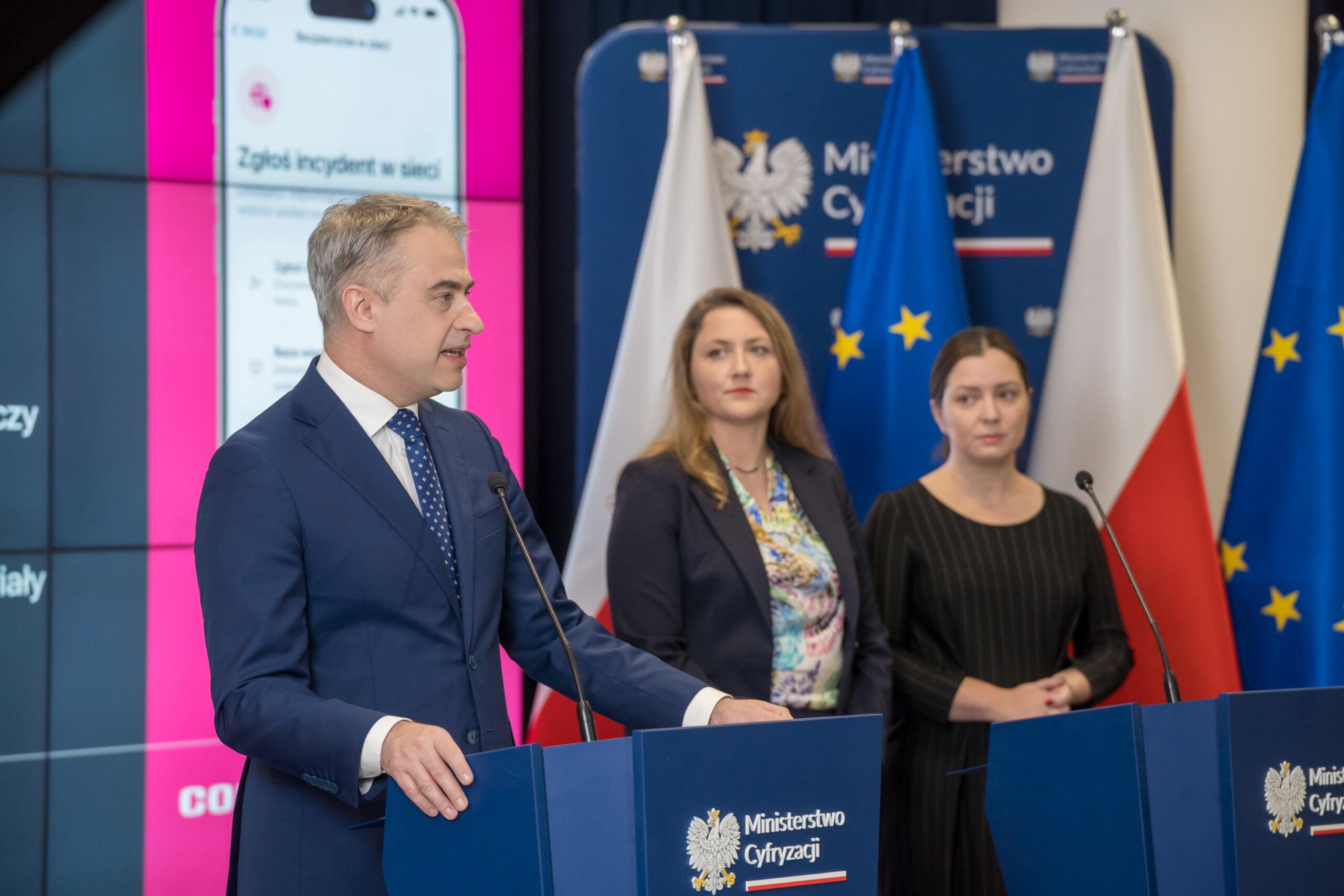 Mężczyzna ( K. Gawkowski) stoi przy mównicy z napisem Ministerstwo cyfryzacji, za nim przy drugiej mównicy dwie kobiety słuchające tego co mówi.