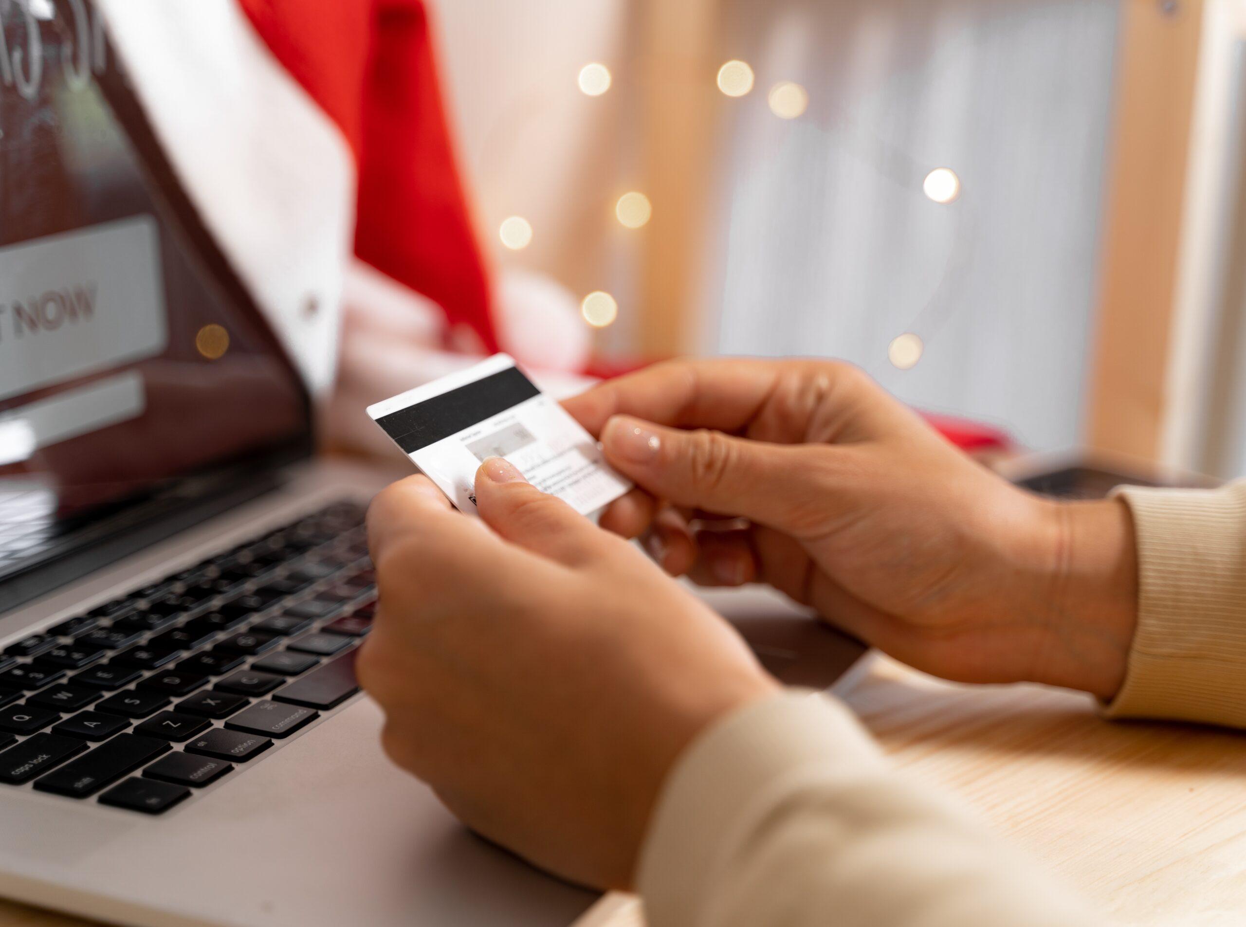 Osoba trzymająca kartę płatniczą w dłoniach przed otwartym laptopem, przygotowując się do zakupów online. W tle rozmyte światełka dekoracyjne, nadające świąteczny nastrój.